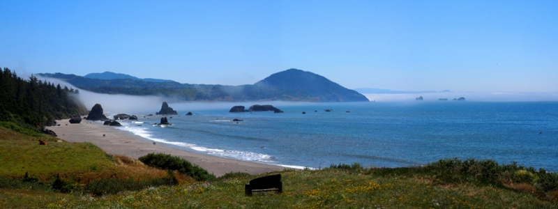 [Two photos stitched together with grass in the foreground, a sandy beach beside it, and then the rocks sticking out of the water near the curve of rock which delineates the bay.]